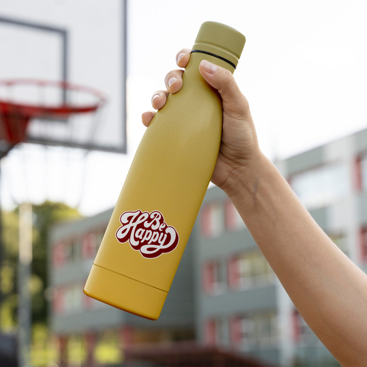 A person holding a yellow Be Happy Sticker water bottle by KosmicSoul in front of a basketball court, highlighting its vibrant color and contrasting against the backdrop of the court.