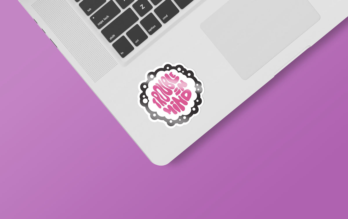 A matte laptop with a Trouble In Mind sticker from KosmicSoul on a purple background.