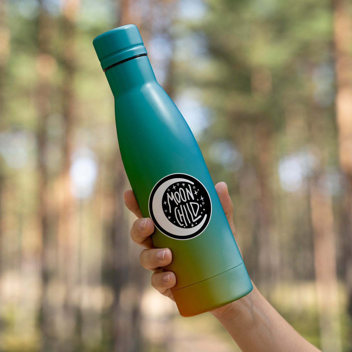 A person holding a Moon Child Sticker by KosmicSoul against the backdrop of a lush forest.