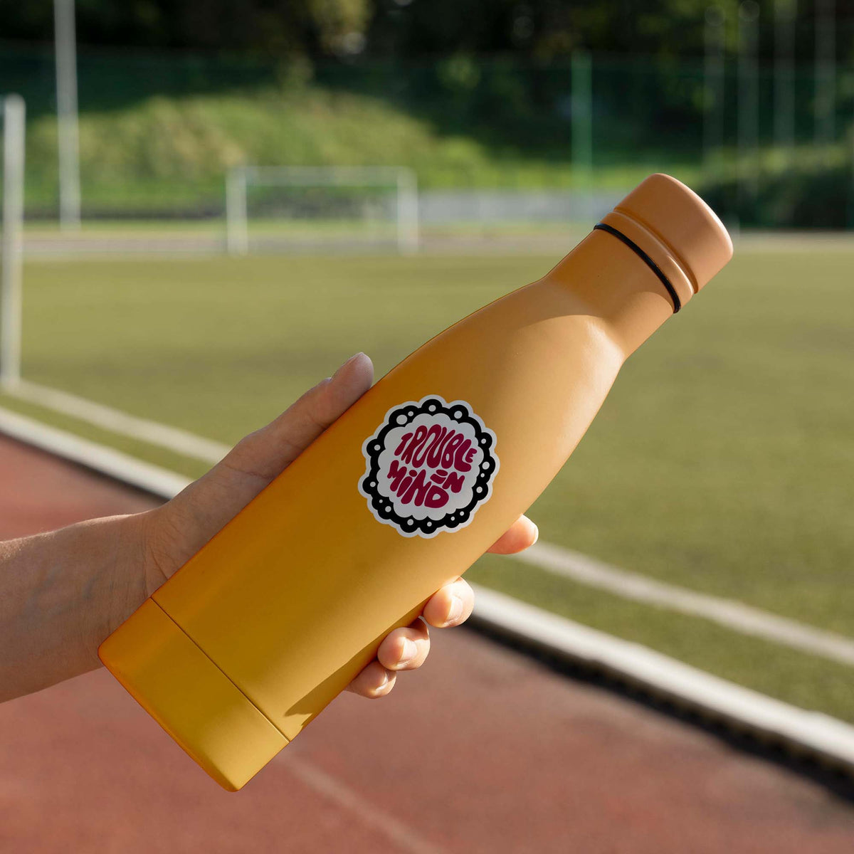 A woman holding a Trouble In Mind Sticker with a matte finish in front of a soccer field.