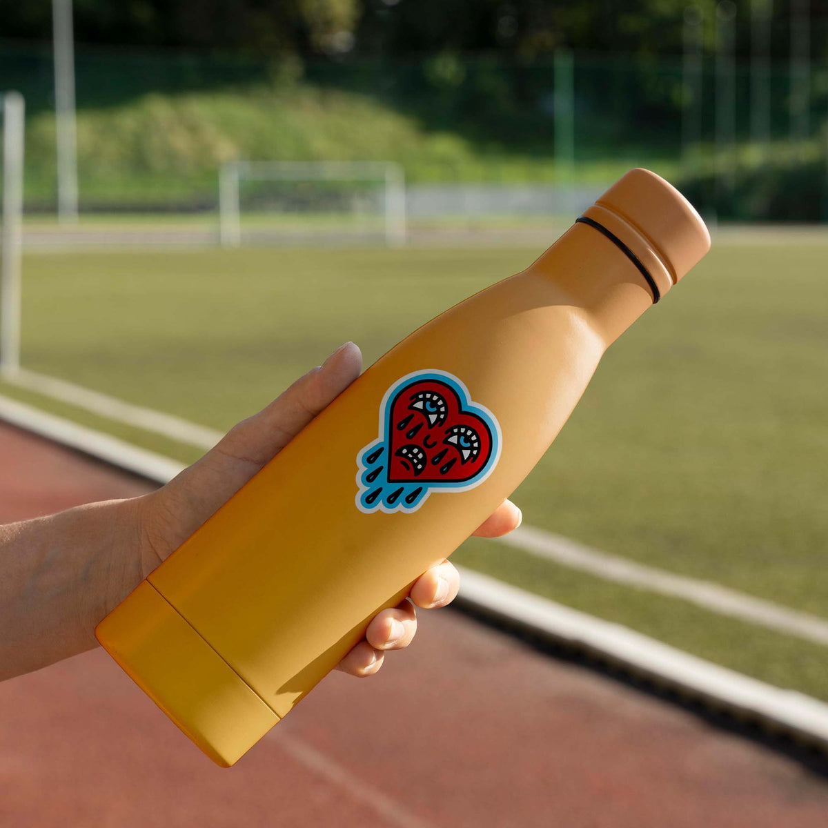 A person holding a yellow KosmicSoul water bottle with a Crying Heart sticker on it that has a matte finish.